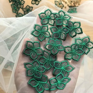 May include: Close-up of several dark green floral appliques. The appliques are in the shape of five-petal flowers with detailed stitching. They are arranged on a sheer white fabric, with some appliques scattered in the background.