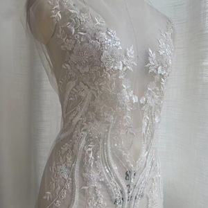 May include: White lace fabric with floral and leaf designs, embellished with sequins. The lace is delicate and intricate, and would be perfect for a wedding dress or other special occasion garment.