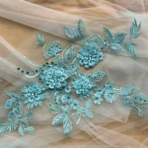 Puede incluir: Primer plano de una aplicación floral azul claro con lentejuelas y cuentas sobre una tela blanca transparente. La aplicación presenta un grupo de flores y hojas.