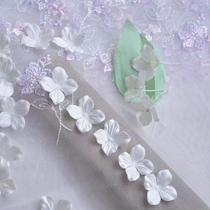 Op de afbeelding: Close-up van witte stof met delicate, driedimensionale witte bloemversieringen. De stof heeft een doorschijnend, netachtig uiterlijk, met geborduurde lavendelkleurige bloemaccenten. Een lichtgroen blad met bevestigde witte bloemen is ook aanwezig.
