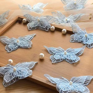 May include: Light blue lace butterfly appliqués with pearl centers are arranged on a wooden tray. The delicate butterfly wings are outlined in white thread. Several white pearls are scattered around the butterflies.