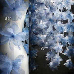 Puede incluir: Una exhibición de flores de tela azul claro, algunas en cascada, otras unidas a una forma blanca. Las flores tienen pétalos delicados y en capas. La imagen incluye texto en la esquina inferior izquierda.