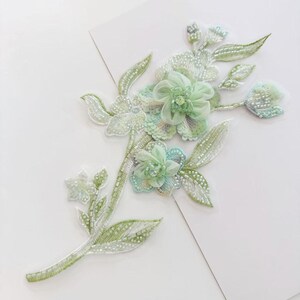 May include: A decorative floral applique featuring light green and mint-colored fabric petals, intricate beadwork, and sequins arranged in a branch-like pattern. The design includes layered blossoms, detailed leaves, and stems crafted with shimmering beads.