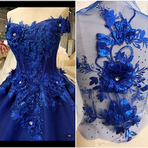 Può includere: Un abito blu reale con spalle scoperte con abbellimenti floreali e accenti scintillanti. Il corpetto presenta un ricamo floreale intricato e una gonna ampia. La schiena dell'abito mostra un'applicazione floreale dettagliata con paillettes e un gioiello centrale.