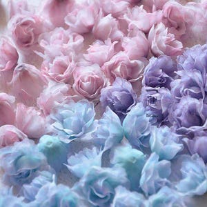 May include: An array of delicate, handmade fabric flowers in shades of pink, lavender, and blue. The flowers are arranged closely together, creating a soft, textured surface. Ideal for crafting or decorative projects.