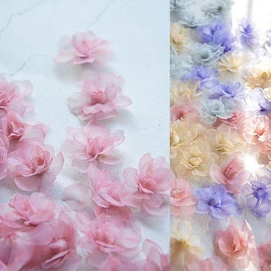 Peut inclure: Gros plan sur des fleurs délicates en tissu translucide dans des tons de rose, pêche et lavande. Les fleurs sont dispersées sur une surface blanche, créant une esthétique douce et aérienne. Idéal pour l'artisanat ou la décoration.