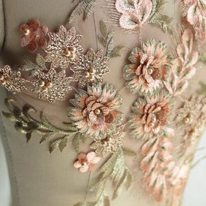 May include: A close-up of a delicate floral lace applique with 3D flowers in shades of peach and green. The applique is adorned with small pearl beads and is perfect for adding a touch of elegance to clothing or crafts.