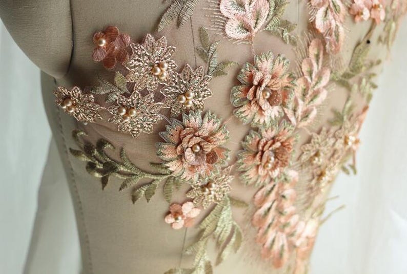 May include: A close-up of a delicate floral lace applique with 3D flowers in shades of peach and green. The applique is adorned with small pearl beads and is perfect for adding a touch of elegance to clothing or crafts.