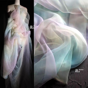 Puede incluir: Una tela fluida en tonos pastel de rosa, azul y amarillo. El material transparente está drapeado sobre un maniquí, con un primer plano de la textura de la tela a la derecha. La tela tiene una calidad suave y etérea.