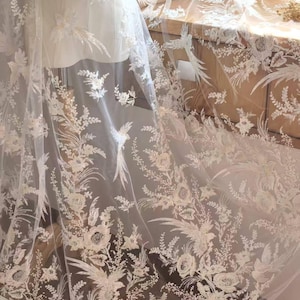 May include: A sheer white tulle fabric featuring intricate embroidery of floral patterns and bird motifs. The delicate design includes detailed blossoms, leafy sprigs, and long-tailed birds scattered across the translucent material in white thread.