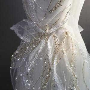 Peut inclure: Une robe en tulle blanc avec un motif délicat et complexe de paillettes dorées et argentées. Les paillettes sont disposées en un motif de feuilles et de fleurs, créant un effet scintillant.
