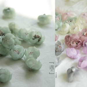 Pode incluir: Close-up de delicadas flores de tecido em cores pastel. As flores são em tons de verde, rosa e roxo, com detalhes intrincados. A imagem inclui texto vertical.