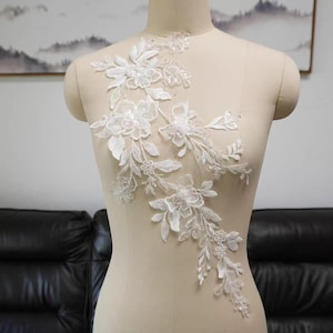 Könnte beinhalten: Weiße florale Spitzenapplikation mit detaillierten Blumen- und Blattmotiven. Die Applikation wird auf einer Schaufensterpuppe präsentiert, die ihre zarte Textur und komplizierten Muster zeigt. Ideal für Brautmode oder Bastelprojekte.
