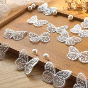 May include: Delicate white butterfly appliques with intricate embroidered details are arranged on a wooden tray, alongside scattered faux pearls. The butterflies vary in size and are ideal for crafting and embellishments.