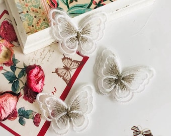 May include: Three white lace butterfly appliques with gold glitter accents. The butterflies are layered on top of each other and are arranged on a white surface.