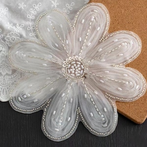 Puede incluir: Aplicación floral blanca con un diseño de pétalos en capas. Los pétalos están delineados con pequeñas cuentas blancas y presentan un adorno central de cuentas. La aplicación está hecha de una tela fina y delicada.