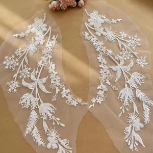 Può includere: Due applicazioni in pizzo bianco simmetriche con intricati ricami floreali e botanici su una base in rete trasparente. Il design presenta fiori dettagliati, fiorellini a forma di stella e rametti frondosi disposti a cascata.
