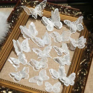 May include: A collection of white lace butterfly appliques with delicate details, some adorned with small pearls. The butterflies are arranged on a neutral-toned surface within a decorative gold frame, creating a whimsical and elegant aesthetic.