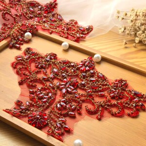 May include: Close-up of red and gold beaded lace appliques with a floral design. The appliques are embellished with red rhinestones and gold beads. Three white pearls are scattered on a wooden tray.