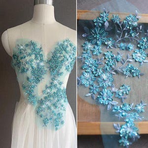 May include: A sheer bodice overlay featuring intricate turquoise floral embroidery with sparkling sequins and beads. The design showcases a dense cluster of blossoms and leaves on a fine mesh fabric, displayed on a dress form and as a flat piece.