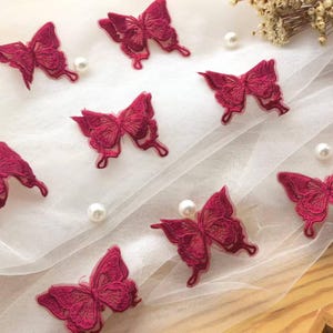 Può includere: Una collezione di toppe ricamate a forma di farfalla color magenta intenso, disposte su un tessuto bianco semitrasparente. Ogni toppa presenta intricati dettagli in filo a forma di ali di farfalla, accompagnati da piccole perle bianche.