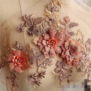 May include: A close-up of a delicate floral applique with intricate details. The applique features a combination of gold and peach-colored flowers and leaves, with small pearl-like beads scattered throughout. The applique is perfect for adding a touch of elegance to clothing, accessories, or home decor.