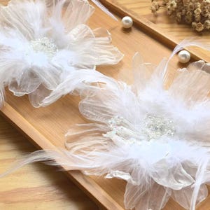 Op de afbeelding: Twee witte florale haaraccessoires met gelaagde, transparante stoffen bloemblaadjes, delicate witte veren en een centraal cluster van heldere, sprankelende kralen. De bloemen zijn naast elkaar geplaatst en hebben een zacht, gelaagd en textuurrijk ontwerp.