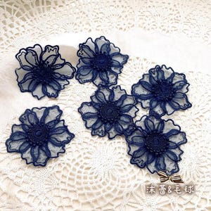 Puede incluir: Seis aplicaciones de encaje de flores azul marino con delicados diseños florales. Las aplicaciones están dispuestas sobre una superficie de crochet blanco.