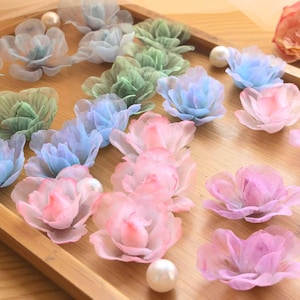 May include: A collection of small, multi-layered fabric flowers in shades of light blue, sage green, soft pink, and lavender. The flowers are arranged on a wooden surface alongside several smooth, round white beads of varying sizes.
