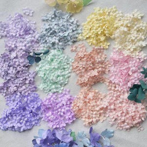 Puede incluir: Un surtido de pequeñas flores de hortensia artificiales en varios colores pastel, incluyendo lavanda, azul, amarillo, melocotón y rosa. Las flores están dispuestas en pequeños montones, creando una exhibición colorida y texturizada.