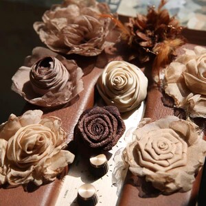 May include: An assortment of handmade fabric flowers in various shades of brown and beige. The flowers are arranged on a brown surface, with some featuring intricate petal designs and others with a more compact, rolled appearance. Some have feather accents.