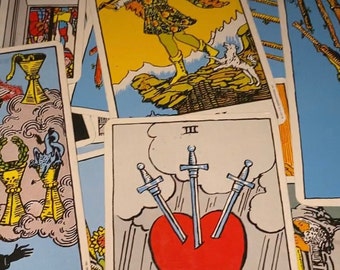 May include: A tarot card reading with a spread of cards, including the Three of Swords card, which features a red heart pierced by three swords. The card is surrounded by other cards, including the Fool card, which depicts a jester-like figure standing on a rock.