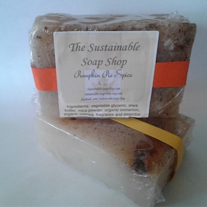 Könnte beinhalten: Zwei Seifenstücke "Pumpkin Pie Spice" in klarer Plastikfolie verpackt. Die Seife ist hellbraun mit sichtbaren Sprenkeln. Ein Etikett trägt die Aufschrift "The Sustainable Soap Shop" und listet die Zutaten auf. Ein orangefarbenes und gelbes Band umwickelt die Seife.