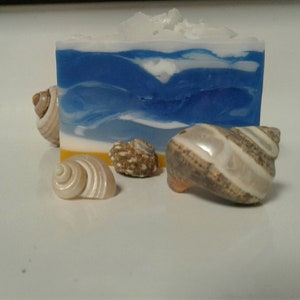 May include: A blue and white layered soap bar with a white top and a blue and white wave design. The soap bar is sitting on a white surface with four seashells.