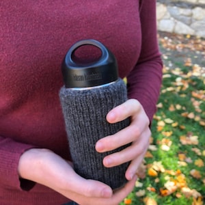 May include: A black stainless steel water bottle with a grey knitted cosy. The bottle has a black lid with a carrying handle. The brand name "Mean Green" is visible on the bottle.