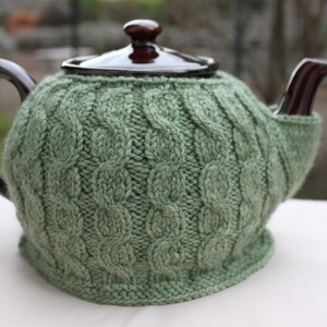 May include: A green knitted teapot cozy with a cable knit pattern. The cozy is on a brown teapot with a black handle and spout.