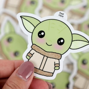 May include: A selection of stickers depicting a cartoon character with large eyes, green skin, and large ears. The character is wearing a tan coat with a brown fur-like collar. The stickers have a white border and the text "Peel Here".