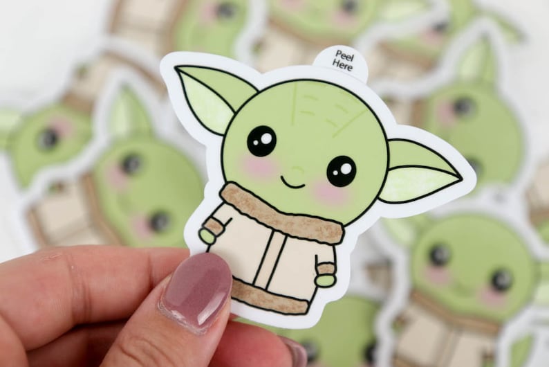 May include: A selection of stickers depicting a cartoon character with large eyes, green skin, and large ears. The character is wearing a tan coat with a brown fur-like collar. The stickers have a white border and the text "Peel Here".