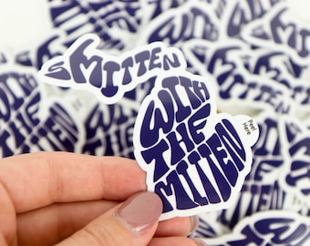 May include: A blue and white sticker shaped like the state of Michigan with the text "With the Mitten" in a stylised font. The sticker has a peel-off backing.