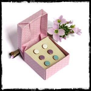 May include: A pink origami box with a beige interior, containing six pairs of small round stud earrings. The earrings are in three colours: white, purple, and blue. The box is open and a sprig of pink flowers is placed next to it.