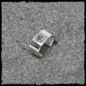 May include: A silver ring featuring a blue gemstone set in the centre. The ring is crafted from hammered metal and has a simple, modern design.