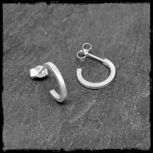 May include: A pair of silver hoop earrings with a simple, minimalist design. The earrings are made of a smooth, brushed metal and have a slightly curved shape.
