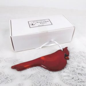 May include: A glossy red cardinal ornament with a white ribbon, on a white lace surface. A white box with the text "Wayne Village Pottery" is in the background.