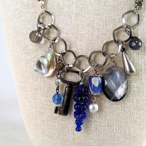 Puede incluir: Un collar plateado con una variedad de colgantes, incluyendo una llave, cuentas de vidrio azul y piedras iridiscentes. El collar tiene una cadena de eslabones entrelazados, sobre un fondo neutro.