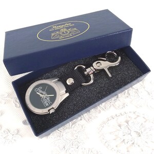Puede incluir: Un juego de reloj y llavero presentado en una caja azul marino. El reloj tiene una esfera negra con manecillas plateadas y una caja plateada. El llavero es plateado con una correa de cuero negro. La tapa de la caja tiene texto dorado.