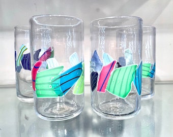 May include: Four clear glass tumblers with a colorful geometric design. The design features green, blue, pink, and white shapes.