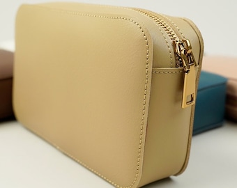 Può includere: Una pochette in pelle beige con cerniera e tiretto dorato. La pochette è appoggiata su una superficie bianca.