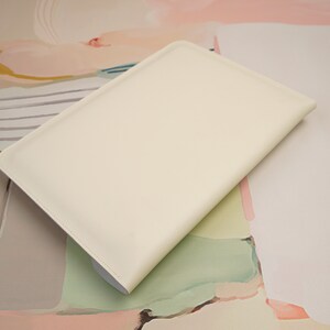 May include: A white leather laptop sleeve with a smooth, matte finish. The sleeve is lying on a colorful abstract background.