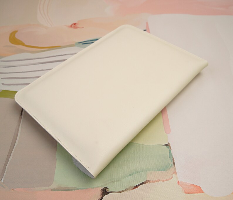 May include: A white leather laptop sleeve with a smooth, matte finish. The sleeve is lying on a colorful abstract background.