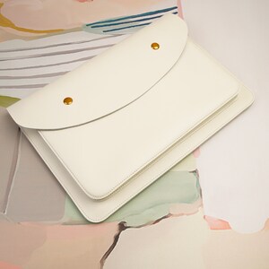 May include: A white leather envelope clutch with a flap closure and gold accents. The clutch is lying on a textured surface with abstract patterns in shades of pink, yellow, and blue.